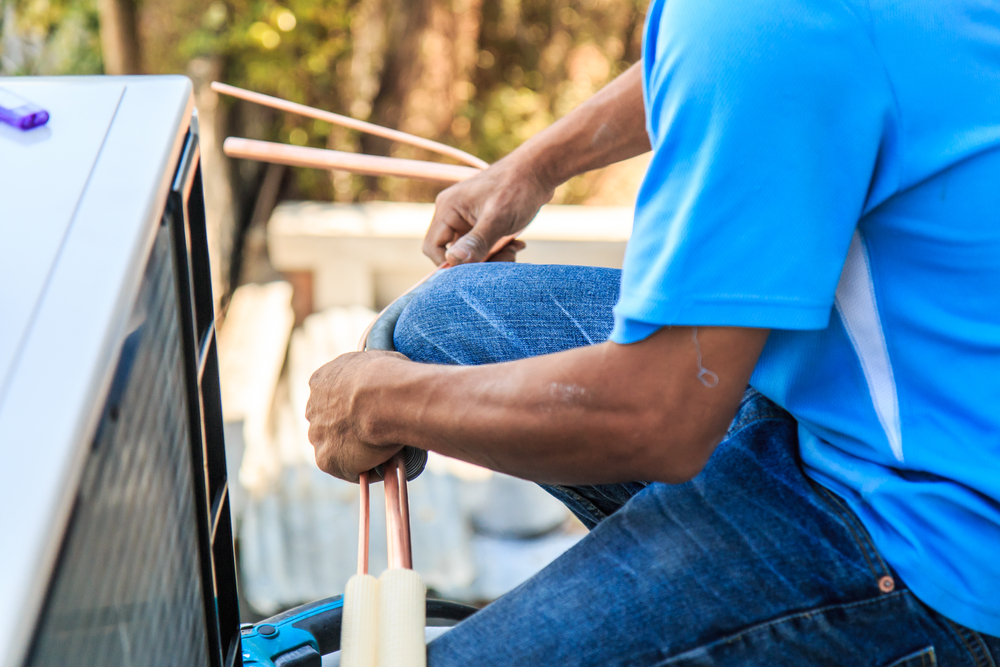 A man in blue jeans and a blue shirt servicing an outdoor air conditioner unit.